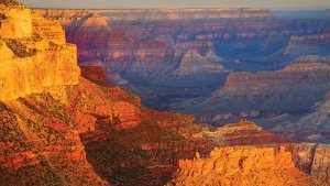 A stunning view of the Grand Canyon during sunset showcases the vast expanse of the canyon with its layered reddish-orange rock formations and deep valleys. The light from the setting sun casts a warm glow, highlighting the rugged terrain that plays a crucial role in driving Arizona's economy through tourism.