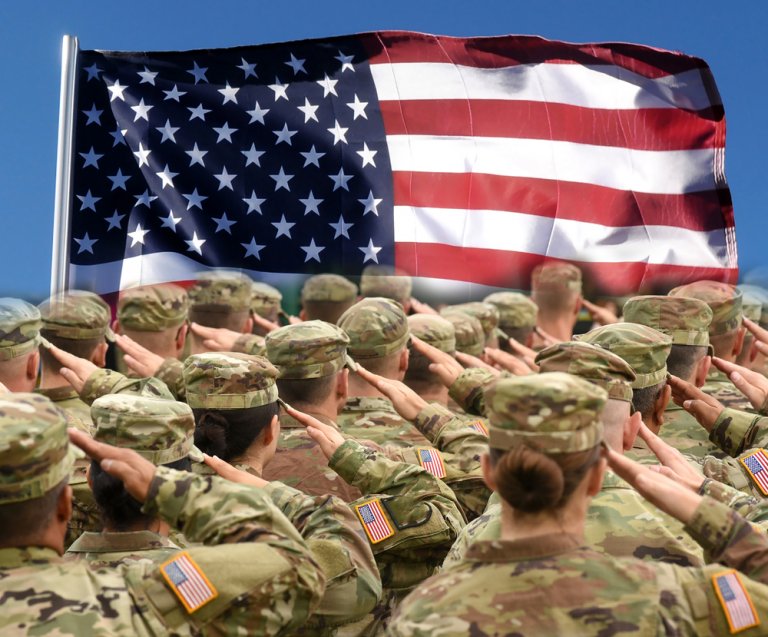 A group of soldiers in camouflage uniforms and helmets stand in formation, saluting a large American flag waving respectfully in the background against a clear blue sky. Some soldiers have American flag patches on their uniforms.