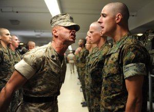 A drill instructor in military uniform yells at a line of new recruits standing at attention indoors. All recruits have shaved heads and are dressed in camouflage uniforms, holding stoic expressions. The setting, resonating with a sense of respect and discipline, appears to be a military training facility.