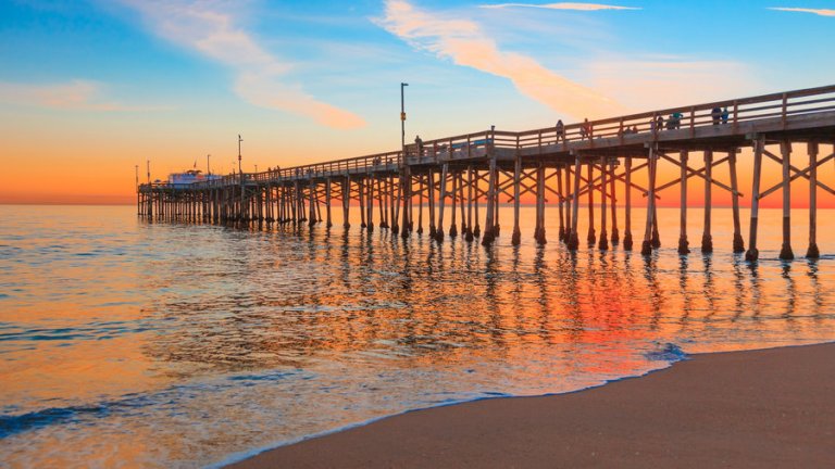 A long wooden pier extends over the calm California ocean water at sunset. The sky is painted in vibrant hues of orange, pink, and blue, creating a serene atmosphere. The reflection of the pier and the sky colors are visible on the water's surface.