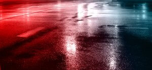A wet street surface glistens under the reflection of red and blue lights at night. The colors blend on the pavement, creating a vibrant, almost metallic effect. The scene is empty, giving a sense of quiet after recent rain, as if waiting for a dispatcher's next directive.