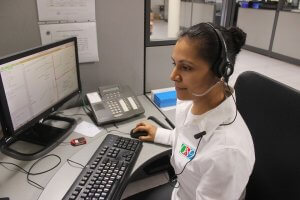 A dispatcher wearing a headset sits at a desk in front of a computer monitor and a telephone. She is typing on a keyboard and appears focused, suggesting she may be providing customer service or technical support. She is dressed in a white shirt with a logo on it.