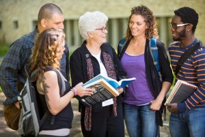 A group of four students stands outdoors, smiling and engaging in conversation with an older woman holding an open book. The students have backpacks and appear to be enjoying the discussion as they communicate eagerly. The background includes a building with large windows.