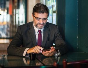 A man with a beard and glasses, dressed in a business suit and red tie, is sitting at a table in a dimly lit cafe. He is looking at his smartphone, perhaps to communicate, while holding a coffee cup on the table. The background features glass panels and a blurred interior.