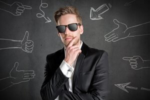 A man in a black suit, white shirt, and sunglasses stands pensively with one hand on his chin. Behind him, a dark gray chalkboard showcases various chalk drawings of hands and arrows pointing in different directions—a great representation of complex decision-making.