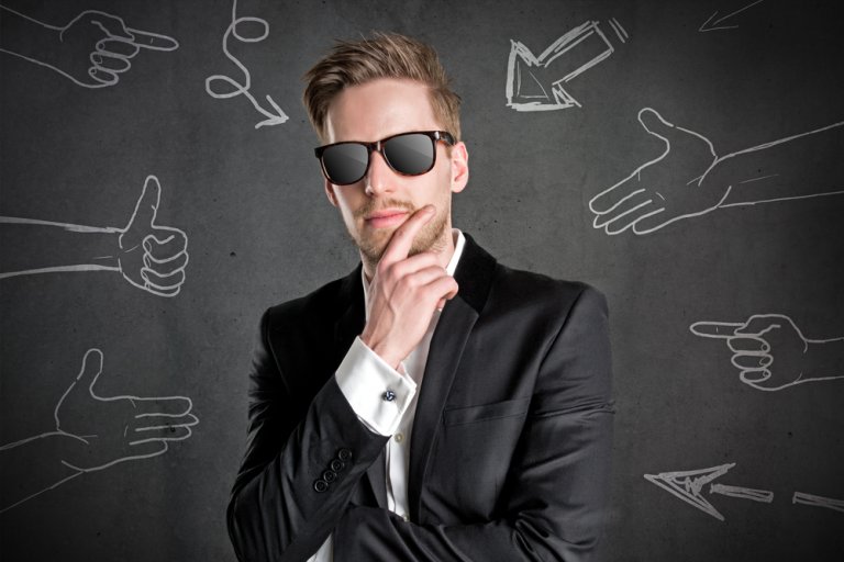 A man in a black suit, white shirt, and sunglasses stands pensively with one hand on his chin. Behind him, a dark gray chalkboard showcases various chalk drawings of hands and arrows pointing in different directions—a great representation of complex decision-making.