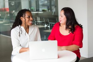 Two women are sitting at a round white table with a laptop in front of them. The woman on the left is wearing a white blouse and has braided hair, while the woman on the right is wearing a red blouse with her dark hair down. They are smiling and engaged in great conversation.