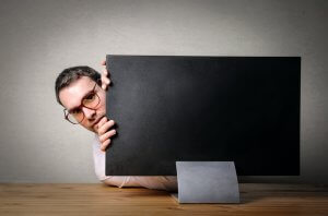 A person with glasses peeks from behind a large computer monitor, likely immersed in a job search. The monitor is on a wooden table, set against a plain, muted wall as the individual looks directly at the camera.