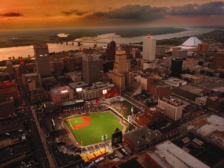 Aerial view of a Memphis cityscape at sunset with a brightly lit baseball stadium at the center. The background showcases a wide river with a bridge, high-rise buildings, and the distant pyramid-shaped structure on the right. The sky is dramatic with orange hues.