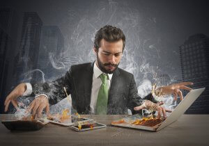 A man in a suit and green tie sits at a desk with a serious expression, frantically working on a smoking, burning laptop. His other devices, including a tablet and phone, are also on fire—clear signs of taking on more than he can handle. The background shows a cityscape with tall buildings.
