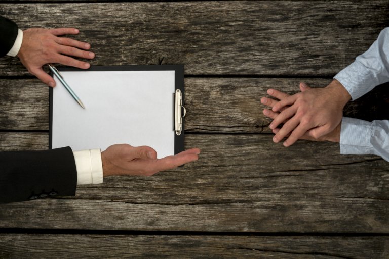 Two people seated across a wooden table with an empty clipboard, a pen, and hands gesturing during a negotiation. One person’s hands are open, indicating an invitation or explanation, while the other's hands are clasped atop the table, possibly indicating contemplation.