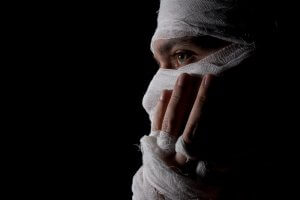 A person with their head and face mostly covered in white gauze bandages, leaving only their eyes partially revealed. Their hand, also wrapped in gauze, gently covers part of their face. The background is completely dark, symbolizing the fragile nature of life.