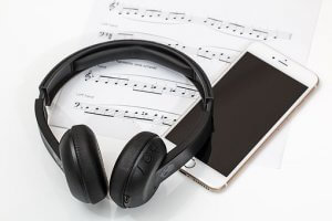 A pair of black over-ear headphones rests on sheets of music notation, bringing the composition to life. Next to the headphones is a white smartphone with a black screen, ready to play what the musical notes and instructions for both hands dictate.
