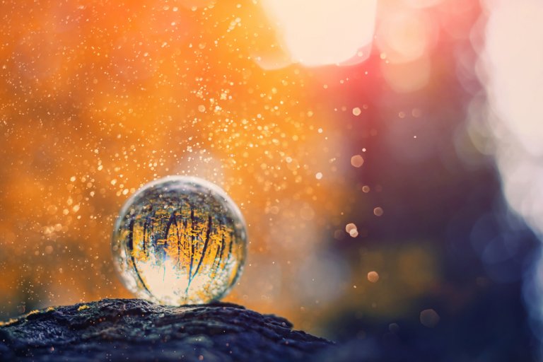 A clear glass sphere rests on a moss-covered rock, reflecting sunlight. The background features a vibrant, bokeh effect with warm hues of orange, red, and yellow, creating a magical atmosphere. Small particles and light flares add to the ethereal ambiance, embodying the beauty of life.