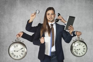 A man in a suit appears to have multiple arms, each holding different objects: a clock in each hand, a magnifying glass, a cup, a phone, and a tablet. This striking image symbolizes multitasking and taking on more responsibilities. The background is plain gray.