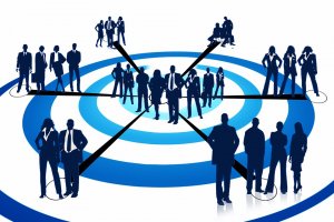 A group of silhouetted business people stands on and around a blue and white target-like graphic. The individuals, taking on more responsibilities, appear in various professional poses, with some standing alone and others in small groups, interconnected by black lines pointing to the center.