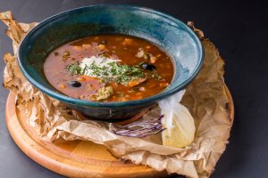 A ceramic blue bowl of hearty soup sits on a wooden board covered with crumpled brown parchment paper. The soup contains various ingredients, including a dollop of sour cream, chopped herbs, and black olives. A wrapped bundle of fresh herbs is placed beside the inviting bowl.