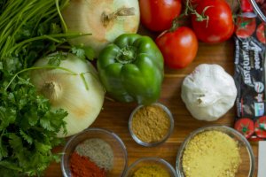 A variety of fresh vegetables and spices are neatly arranged on a wooden surface, perfect for making soup. There are onions, a green bell pepper, tomatoes on a vine, a head of garlic, cilantro, as well as bowls of ground spices and tomato paste in a tube.