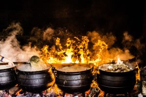 A row of large black pots over an open flame, with bright orange fire and smoke, cooks various ingredients into hearty soup. A person stirs the boiling contents of the leftmost pot with a ladle. The pots, filled to the brim, are being prepared outdoors.