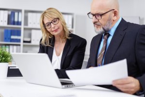 Two business professionals, a woman and a man, are working together at a desk. The blonde woman is looking at a laptop screen, while the bald man with glasses is holding a document. They communicate effectively as they collaborate. Shelves with binders and a plant are in the background.