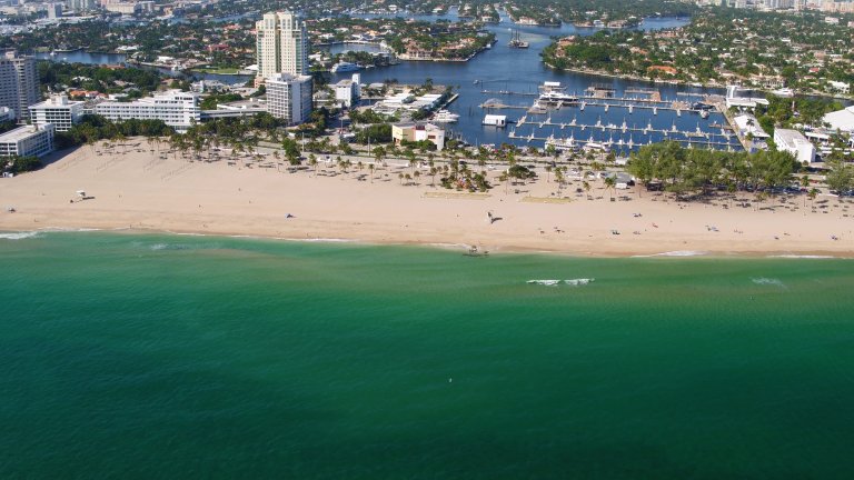 Aerial view of a sandy beach along an ocean with turquoise water. The beach is lined with trees, buildings, and a marina filled with boats. In the background, Fort Lauderdale's cityscape with more buildings and water channels can be seen under a clear blue sky.