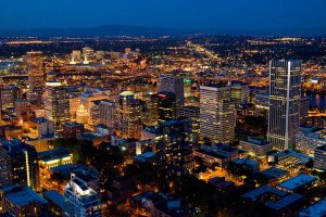 An aerial night view of Portland's cityscape with illuminated buildings, streets, and bridges. The city lights create a vibrant glow, highlighting tall skyscrapers and dense urban infrastructure, set against a dark blue sky and distant, softly lit horizon.