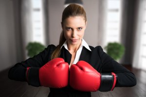 A confident, assertive woman in a black blazer and white shirt stands in a blurred office setting, wearing red boxing gloves and holding them up in a ready position. Her expression is determined, and her hair is pulled back into a ponytail.