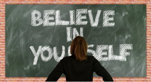 A person with shoulder-length brown hair, seen from behind, stands assertively in front of a chalkboard with the words "BELIEVE IN YOURSELF" written in large white chalk letters. The chalkboard is framed by a brick wall.