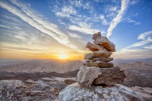 A small stack of balanced rocks in the foreground set against a stunning sunrise or sunset. The sky assertively displays wispy clouds, and rugged mountain terrain stretches out in the background. The colors create a serene and picturesque landscape.