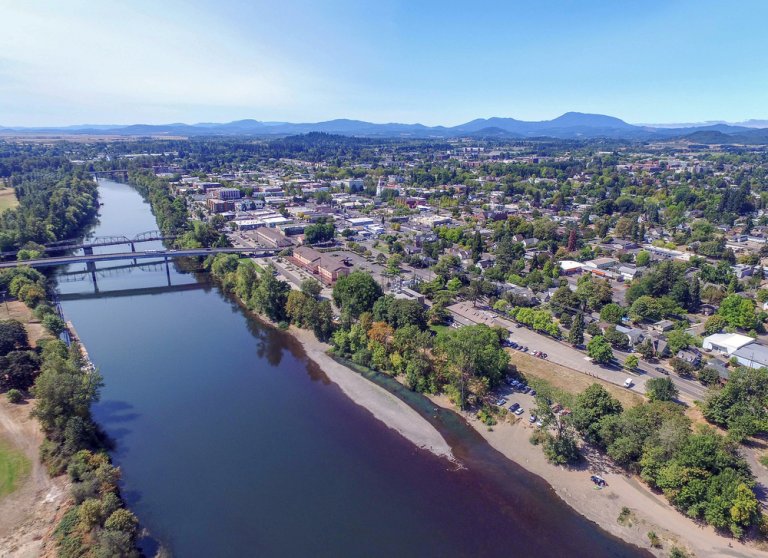 Aerial view of a small city in Benton County alongside a river. A bridge spans the water, connecting two parts of the town. Residential and commercial buildings are intermixed with green spaces and trees. Distant mountains and a clear blue sky form the backdrop.