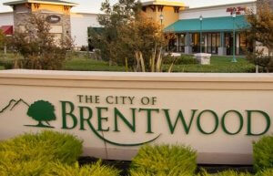 A sign reads "THE CITY OF BRENTWOOD" with a tree and mountain graphic. Behind the sign, various storefronts and trees blend into the commercial area, showcasing well-maintained landscaping under a clear Brentwood sky.