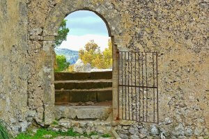 A stone archway with a rusted metal gate stands in an old, weathered wall, leading to a set of steps. Through the arch, trees and mountains are visible under a cloudy sky. The scene exudes a rustic and historical charm, evoking a sense of independence and timeless tranquility.
