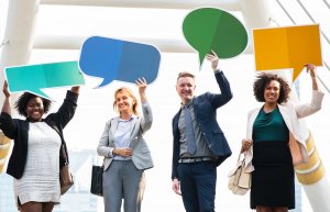 Four diverse professionals stand outdoors holding colorful speech bubble signs above their heads, showcasing engagement. Smiling and dressed in business attire, they stand against a bright architectural backdrop with large beams and a partly cloudy sky.