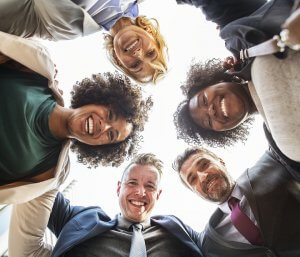 Five people standing in a circle, looking down at the camera with wide smiles, reflecting true engagement. They appear to be colleagues or friends dressed in professional and casual attire, showcasing diverse ages and backgrounds. The bright sky is visible in the background.
