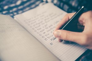 A hand holding a pen is writing in a notebook with a grid-patterned page. The page displays a to-do list with checkboxes, some of which are ticked off, indicating completed tasks. The background is slightly blurred, emphasizing the notebook and hand, much like the focus one would have when updating their resume.
