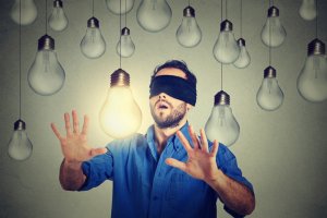 A blindfolded man wearing a blue shirt stands with his hands extended, intuitively touching a lit light bulb among numerous unlit light bulbs hanging around him. The background is a plain gray wall.