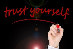 A hand is writing "trust yourself" in red marker on a dark background, reflecting the power of infotuition. Light flares are visible at the center of the image, emphasizing the message. The hand holding the marker appears from the bottom right corner.
