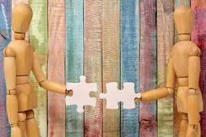 Two wooden mannequins facing each other, each holding a white puzzle piece to connect. The background consists of vertical, multicolored wooden planks. The image conveys themes of collaboration and connection, resembling the essence of infotuition in bringing pieces together seamlessly.