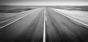 A black and white image captures a straight, empty road starting in the middle and stretching into the horizon. Flanked by flat, barren land, it evokes a sense of vastness and solitude. The sky appears overcast with a hint of a misty atmosphere.