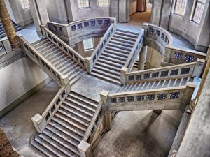 A grand, multi-level stone staircase with intricately designed railings, starting in the middle of the hall, is connected by landings at the corners, forming an elegant, symmetrical pattern. The staircase is illuminated by natural light streaming in through tall, arched windows.