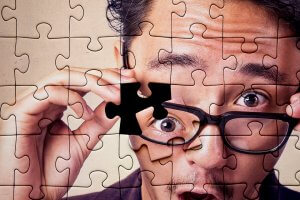 A person with black glasses looks surprised as they hold a missing piece of a jigsaw puzzle in front of their face, starting in the middle. The individual's face is partially covered by the puzzle, with the glasses being a focal point due to the missing piece.