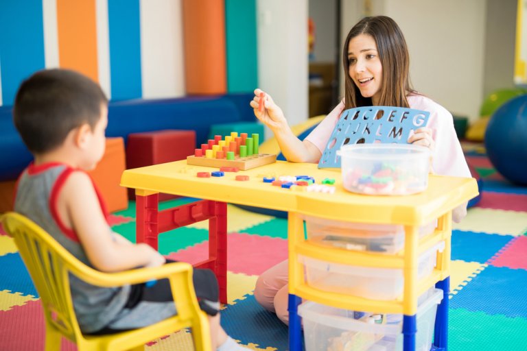 A woman and a young boy are sitting at a table in a colorful room. The woman, specializing in special education, holds a blue chart with letters and uses a pointer while the boy watches. The table features educational toys and tools, and there are soft mats on the floor.