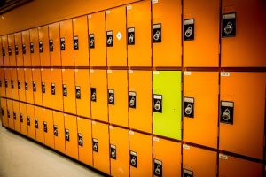 A long row of vibrant orange lockers with black combination locks, one locker stands out as it is painted a bright lime green, creating a visual contrast. The image captures the interior of what seems to be a special education school or gym locker area.
