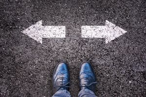 A pair of shiny black dress shoes stand on an asphalt surface, with two white arrows painted on the ground pointing in opposite directions, guiding your path—one arrow to the left and one to the right.