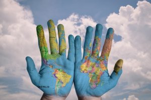 Two hands are held up against a cloudy sky backdrop. The hands are painted with a world map, showing continents and countries in various colors, including North and South America on one hand and Europe, Africa, Asia, and Australia on the other—illustrating a path across the globe.