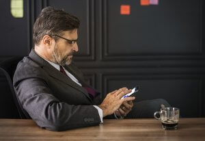 A man in a gray suit and glasses sits at a brown wooden table, clearly engaged in management tasks on his smartphone. The table holds a glass cup with a dark beverage. The background features a dark wall dotted with colorful sticky notes.