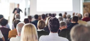 A group of people sits in rows, facing forward, attentively listening to a management expert speaking at the front of the room. The speaker is slightly out of focus, with a large, blank white screen visible behind them.