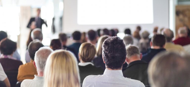 A group of people sits in rows, facing forward, attentively listening to a management expert speaking at the front of the room. The speaker is slightly out of focus, with a large, blank white screen visible behind them.