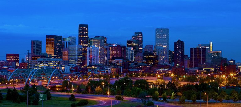 A cityscape of downtown Denver at dusk, showcasing an array of illuminated skyscrapers and buildings. The image captures the bustling energy of the city with light trails from vehicles on the highway in the foreground and a lit bridge crossing over.