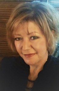 A woman with short, light brown hair and a black blouse smiles warmly at the camera. She has light makeup and green eyes, with a wooden background that evokes the rustic charm of San Marcos behind her.